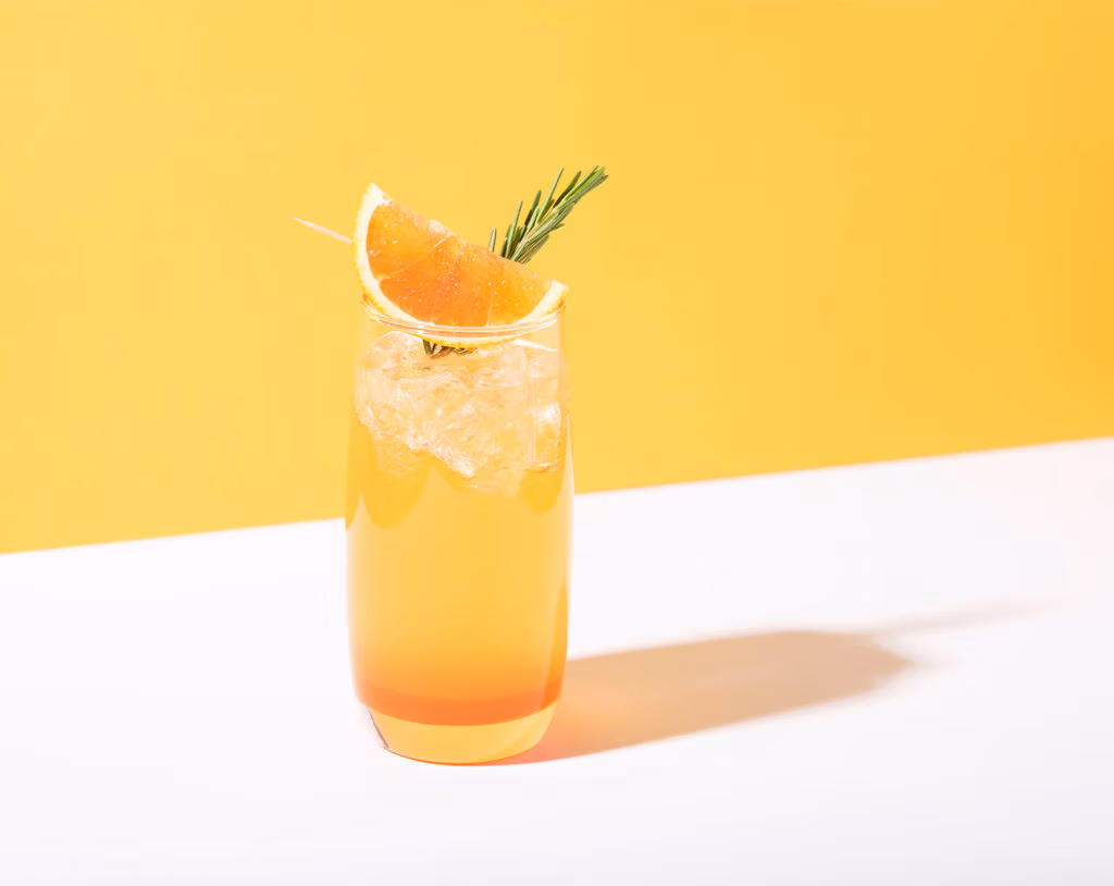 Tall glass of orange cocktail with ice, garnished with an orange slice and a rosemary sprig on a white surface against a bright yellow background.