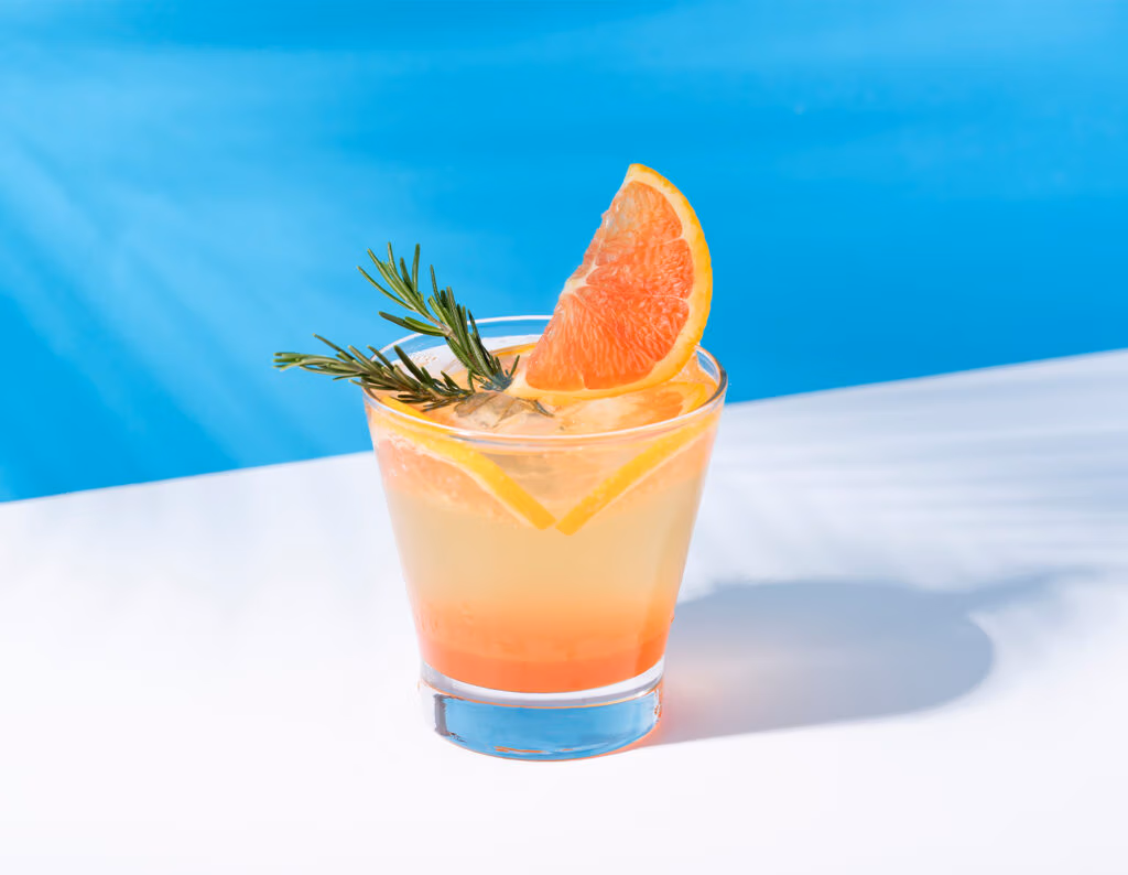 Cocktail in a short glass with orange slice, rosemary sprig, and ice, set against a blue pool background.